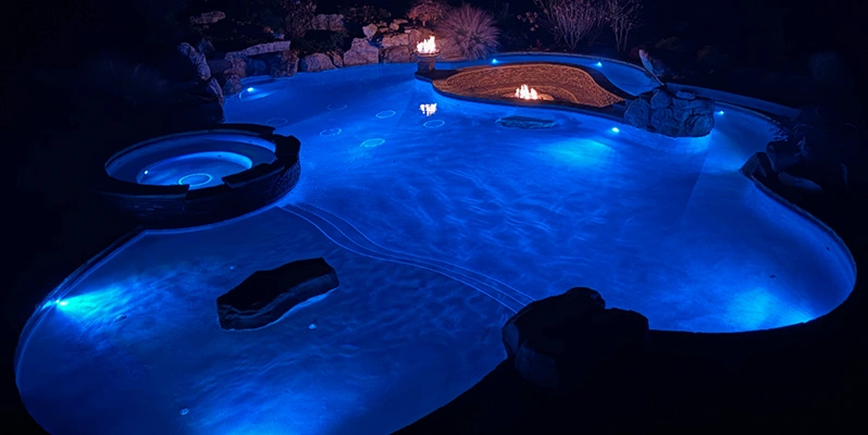 A serene pool illuminated by blue lights, accompanied by a cozy fire pit nearby, creating a relaxing atmosphere.