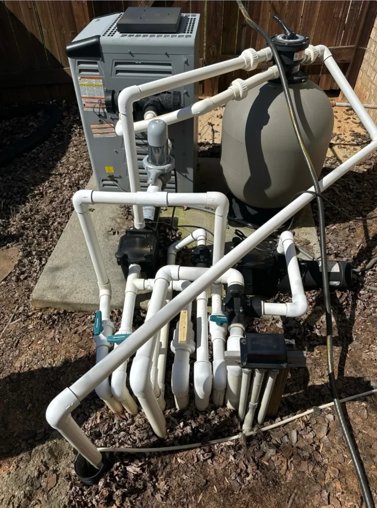 Older pool equipment system with white PVC pipes, pump, and sand filter installed outdoors