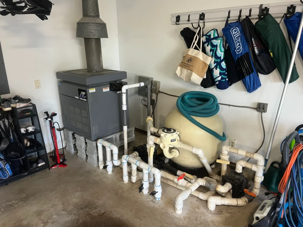 Indoor pool equipment system with heater, sand filter, and PVC piping next to stored gear