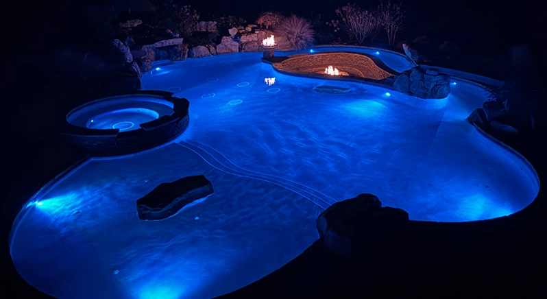 A serene pool illuminated by blue lights, accompanied by a cozy fire pit nearby, creating a relaxing atmosphere.